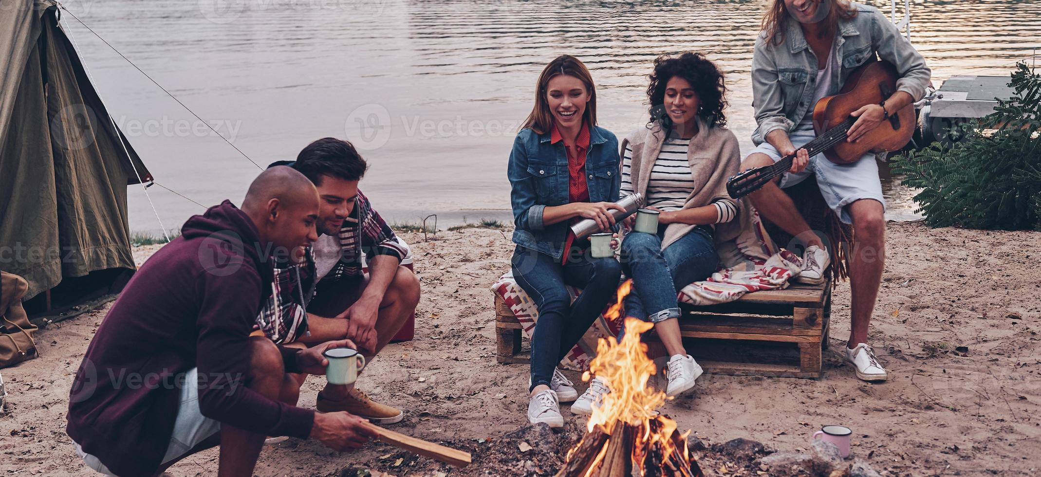 Getting away from it all... Group of young people in casual wear smiling while enjoying beach party near the campfire photo