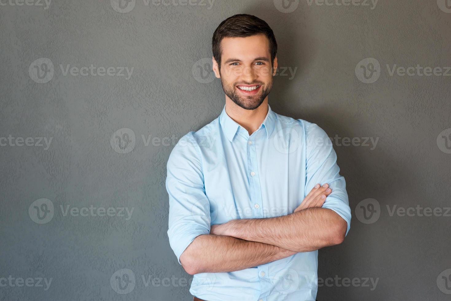 guapo y confiado. un joven guapo con camisa que mantiene los brazos cruzados y sonríe a la cámara mientras se enfrenta a un fondo gris foto