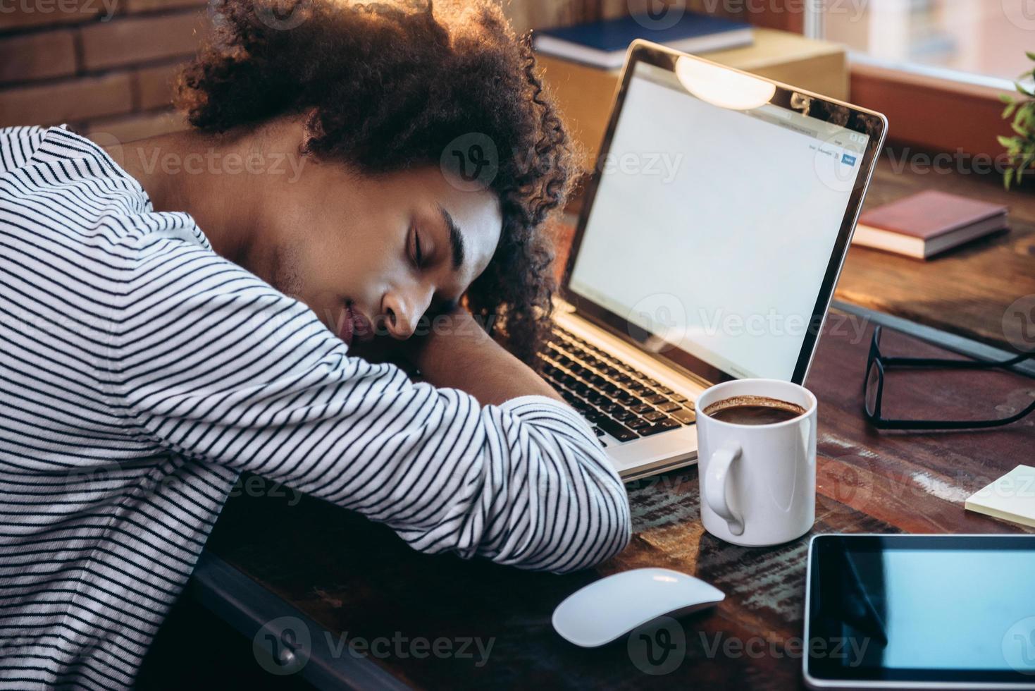 Quick nap. Side view of young African man sleeping on laptop with eyes closed while sitting at his working place photo
