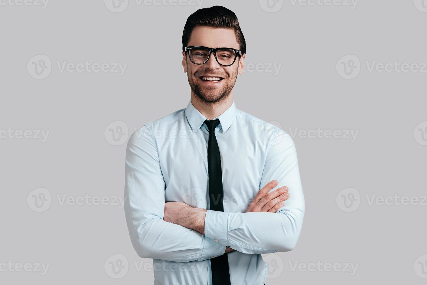 Confident manager. Handsome young man in shirt and tie keeping arms crossed and looking at camera while standing against grey background photo