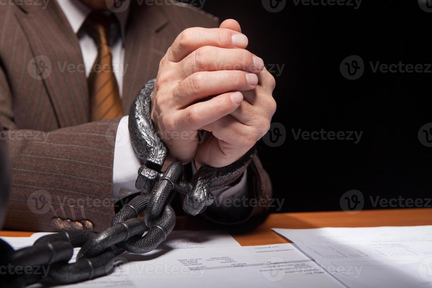 Chained hands. Close-up of man in formalwear with chain on hands sitting at the table and isolated on black background photo