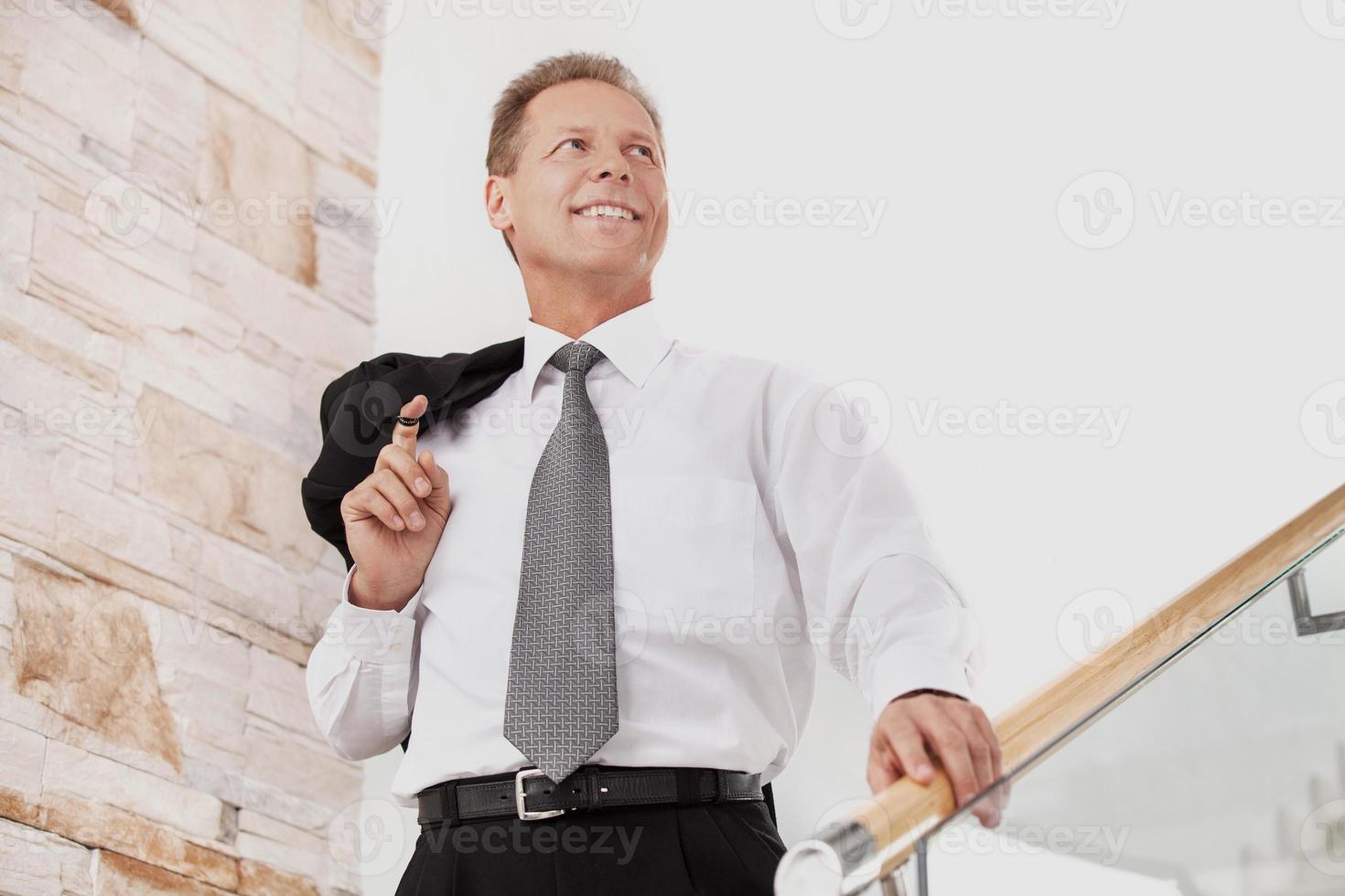 Confident businessman. Cheerful mature man in formalwear smiling while moving down by staircase photo