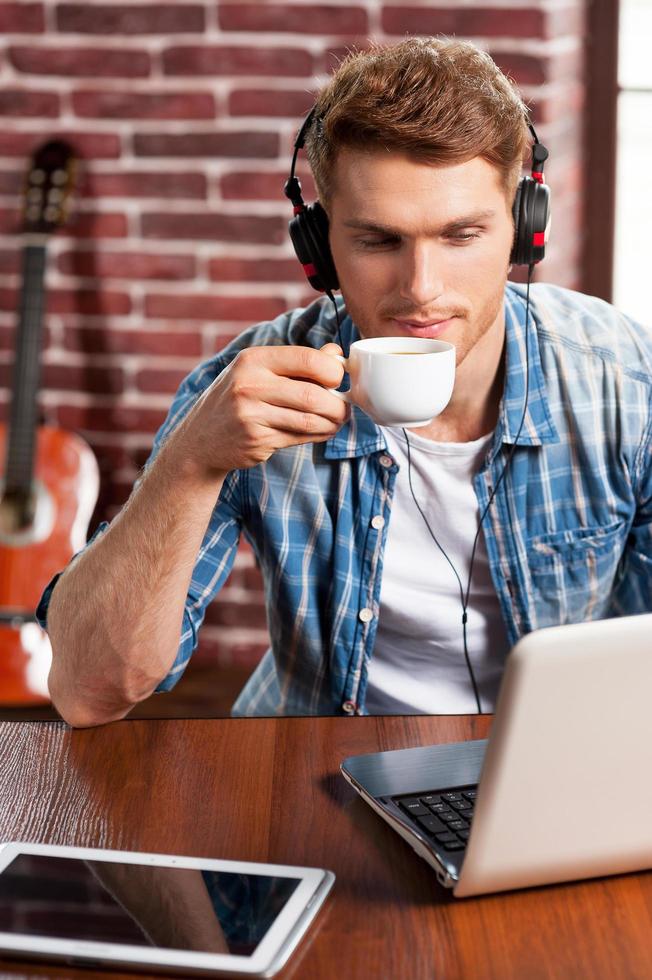 Music is my life. Top view of handsome young man in headphones working on laptop and drinking coffee while acoustic guitar laying in the background photo