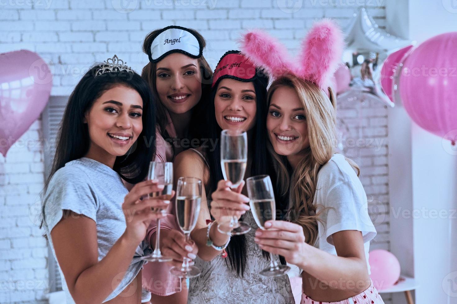 Cheers Four playful young smiling women in pajamas toasting each other while having a slumber party in the bedroom with balloons all over the place photo