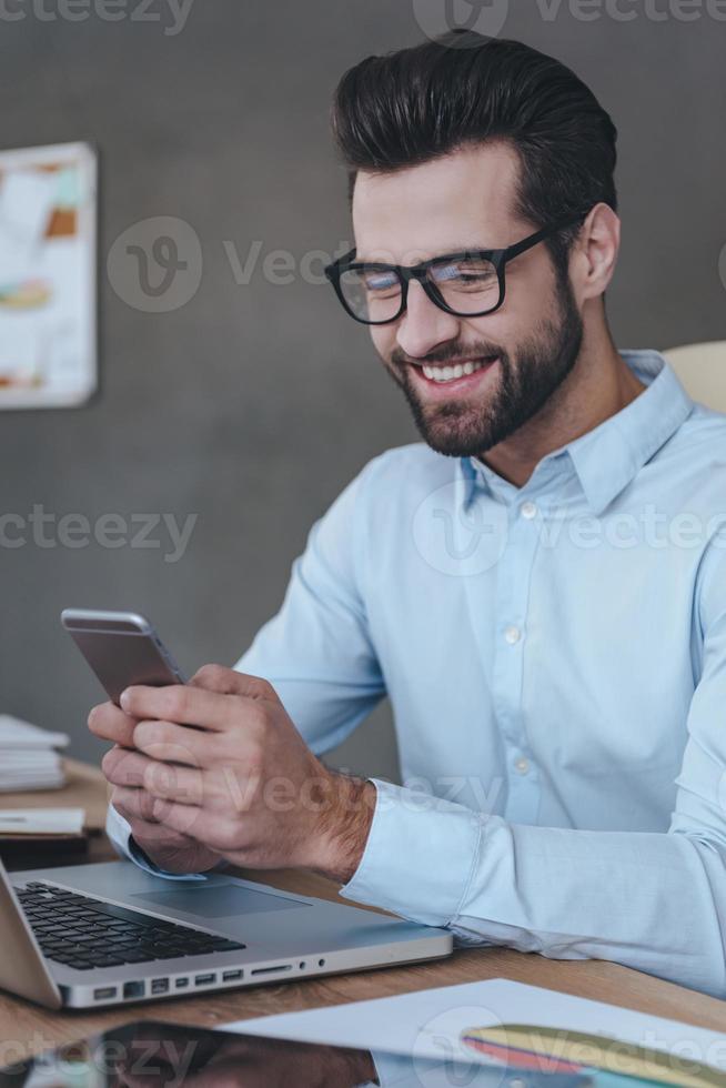 Great news Handsome young man wearing glasses holding smart phone and looking at it with smile while sitting at his working place photo