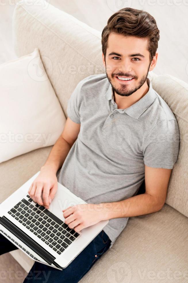 Working at home. Top view of young man working on laptop and looking at camera while sitting on sofa photo