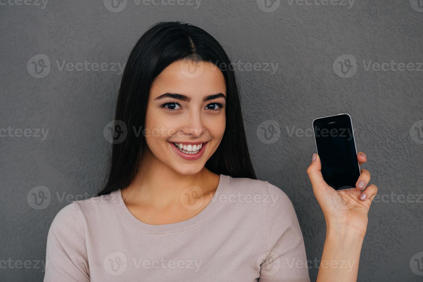 mostrando su nuevo teléfono inteligente. hermosa mujer joven mostrando su teléfono inteligente y sonriendo mientras está de pie contra el fondo gris foto