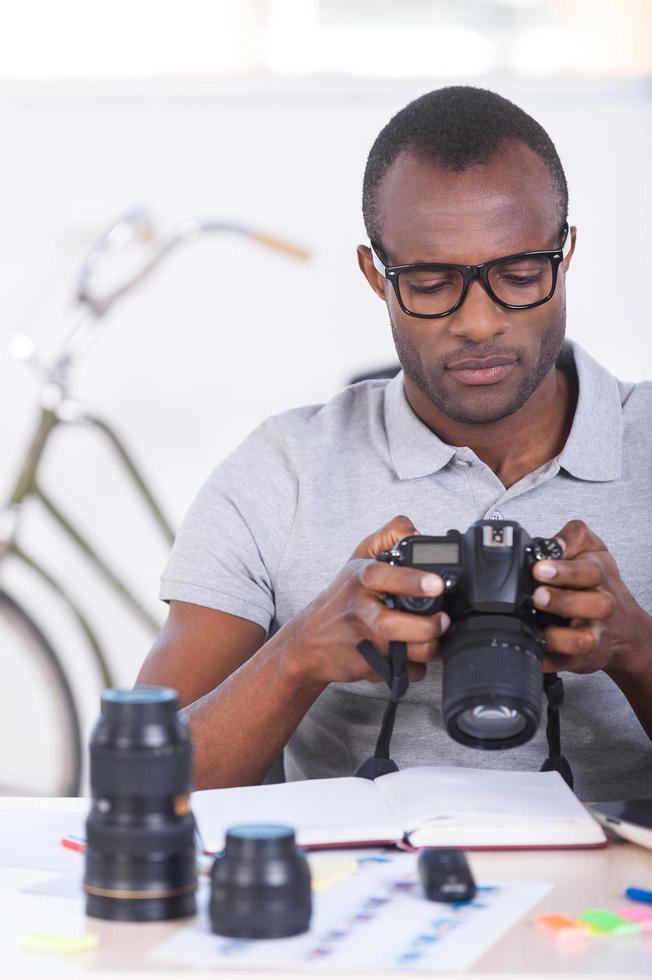 Checking photos. Confident young African man in casual checking photos at his camera while sitting at his working place