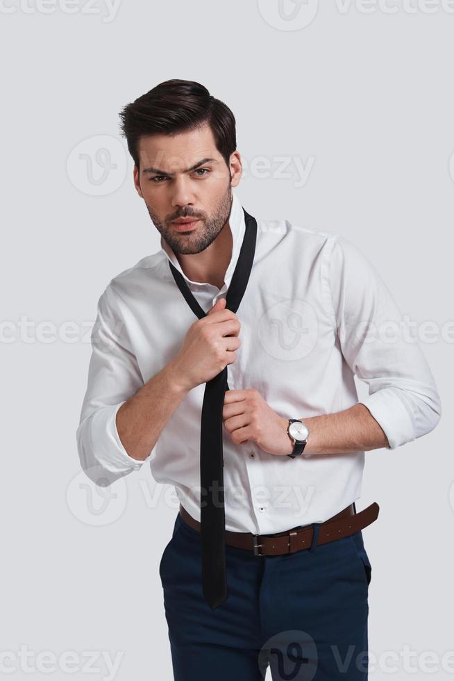 Taking his necktie off.  Tired young man looking at camera and taking on his necktie while standing against grey background photo