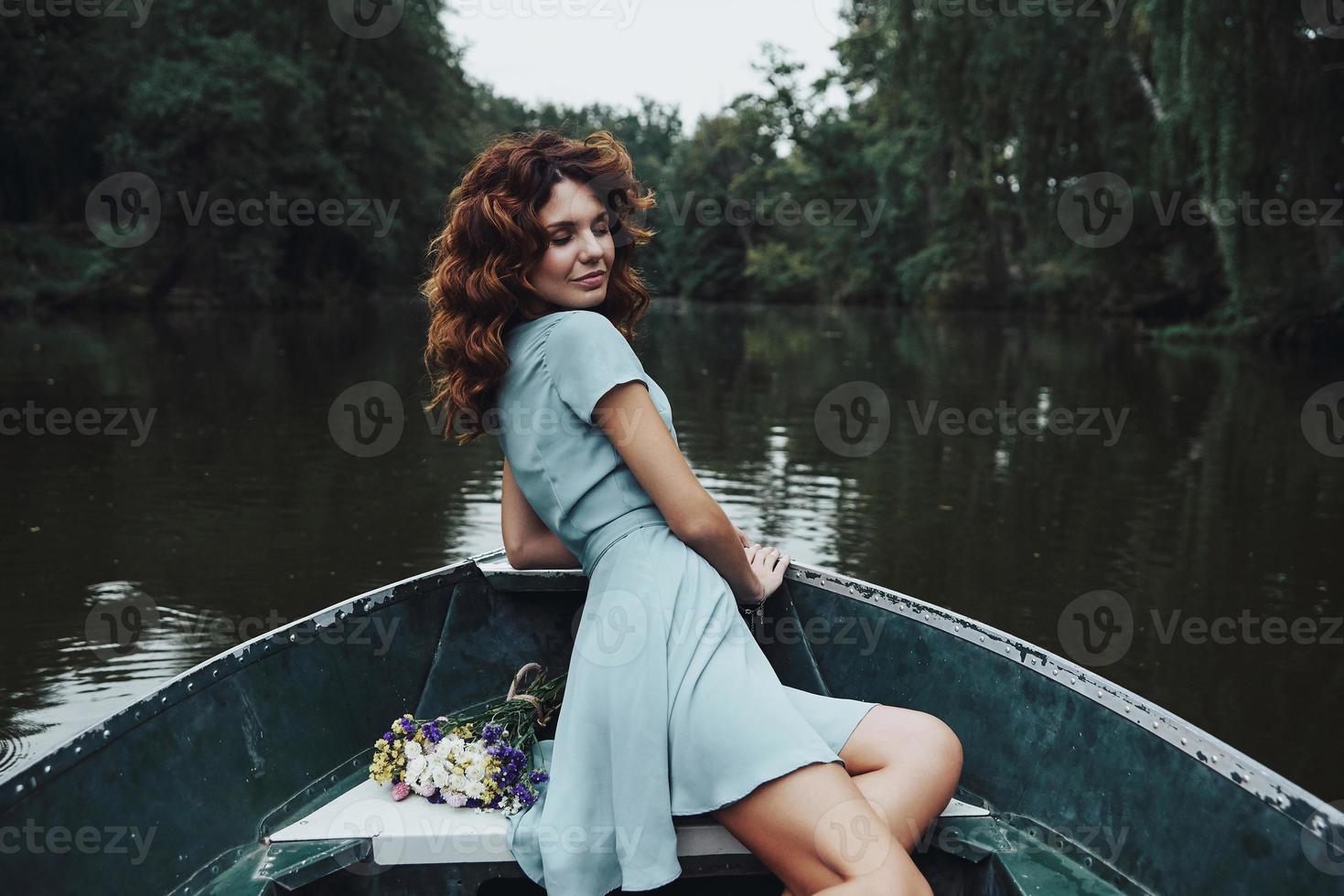 Enjoying carefree day. Beautiful young woman in elegant dress smiling while sitting in the boat photo