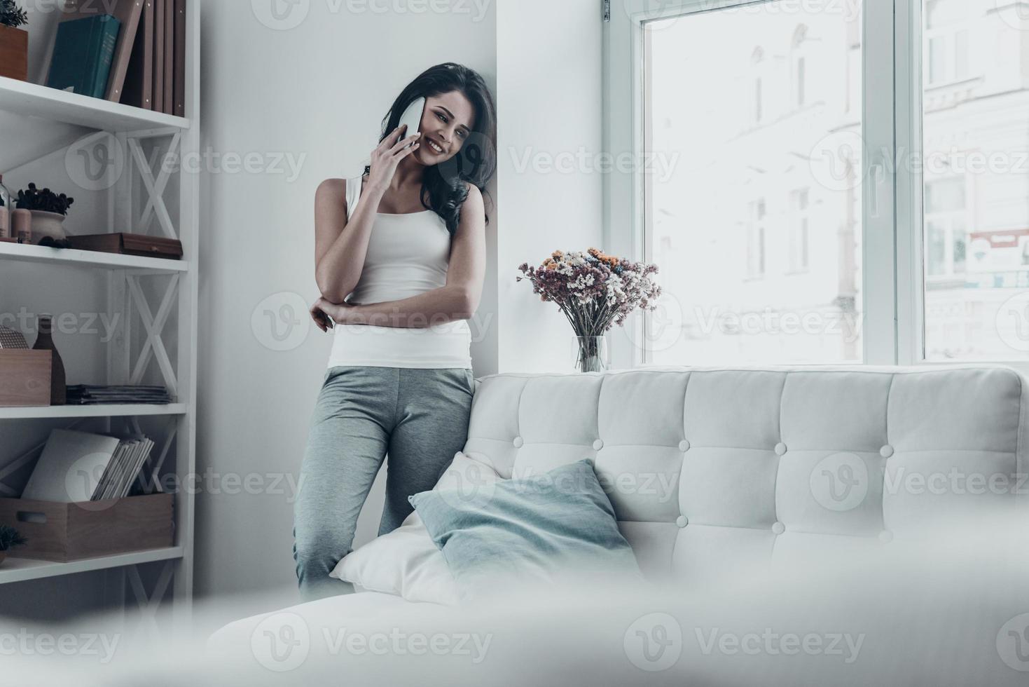 So good to hear from you Attractive young woman talking on the mobile phone and smiling while leaning on the wall at home photo