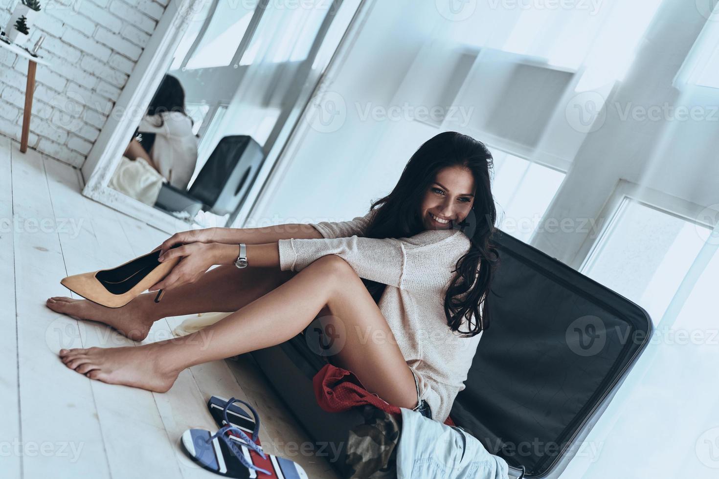 Preparation to vacation.  Beautiful young woman smiling and looking away while sitting on the open suitcase at home photo