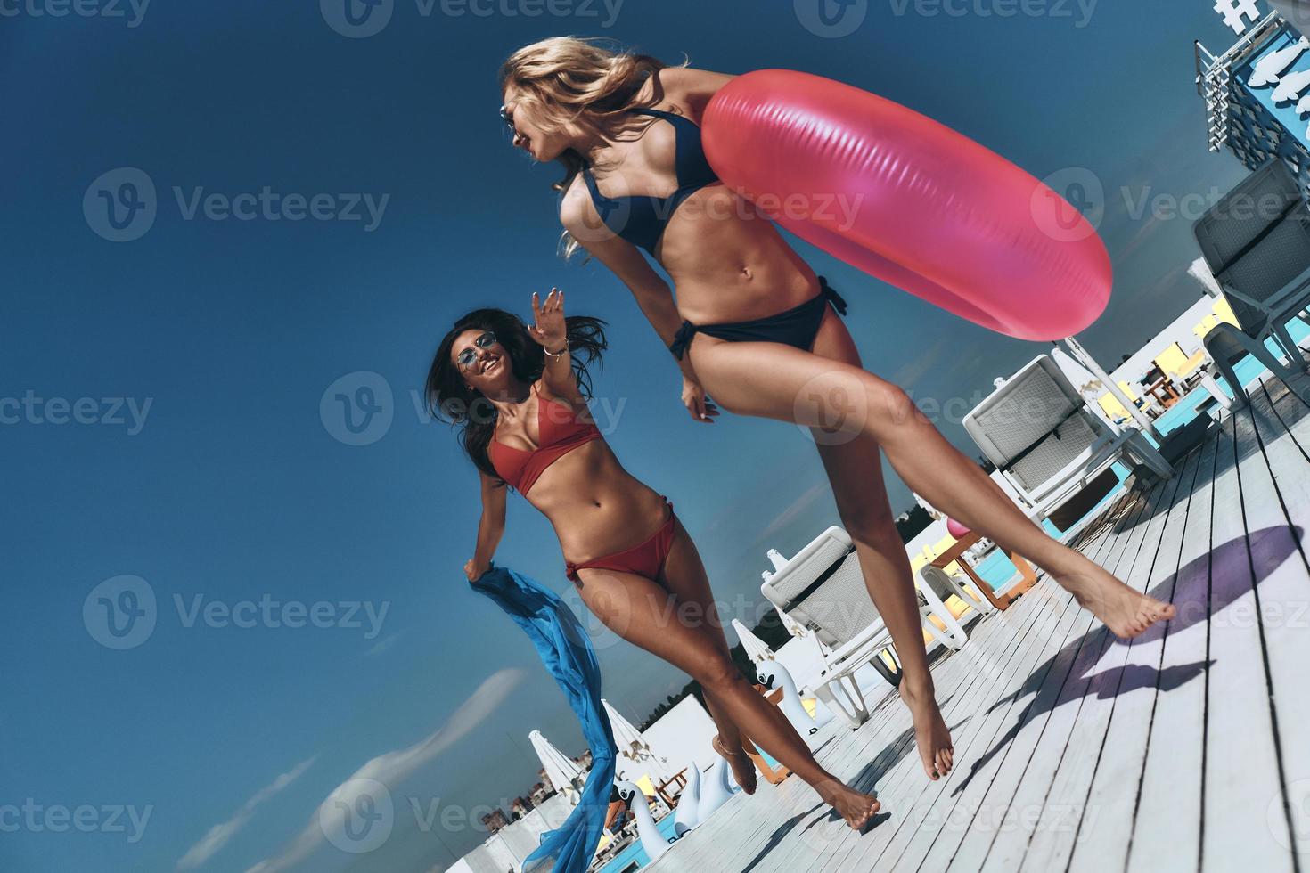 Full length of two beautiful young women in bikini smiling while running near the pool together photo