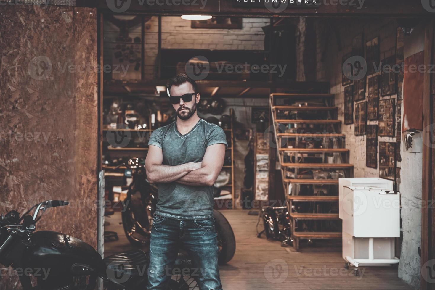 My garage and my rules Confident young man keeping arms crossed and looking at camera while standing near his motorcycle garage or repair shop photo