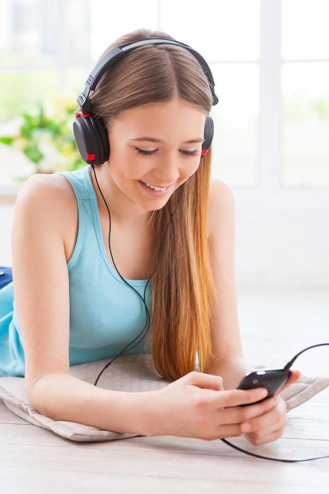 Listening to her favorite music. Cheerful teenage girl in headphones listening to the music while lying down on the floor at her apartment photo