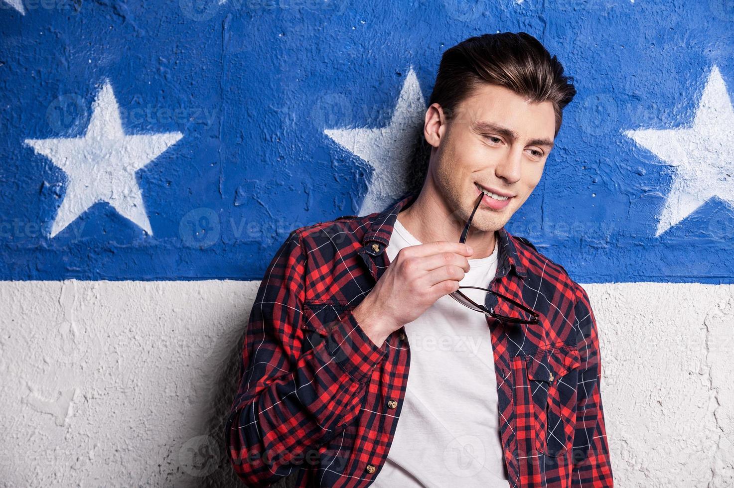 Handsome day dreamer. Cheerful male model in checked shirt holding sunglasses and looking away while standing against background photo