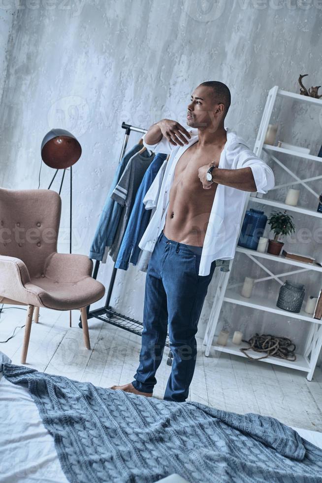 Preparing for the big day. Handsome young African man putting on shirt while standing in the bedroom at home photo