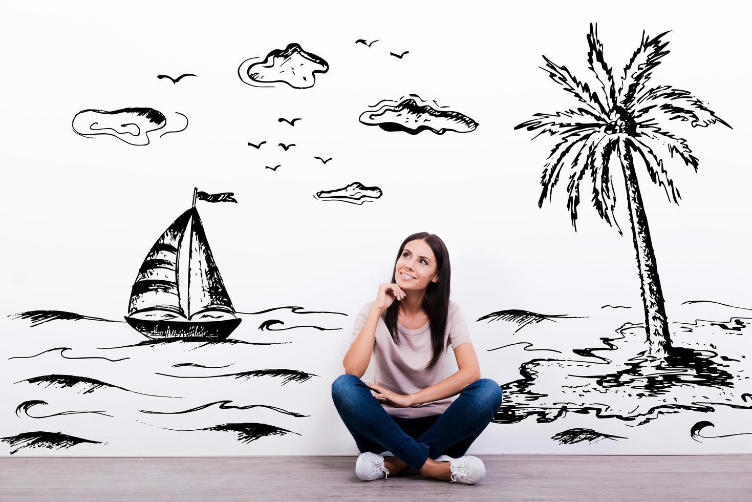 Dreaming about vacation. Cheerful young woman smiling while sitting on the floor with illustration in the background photo