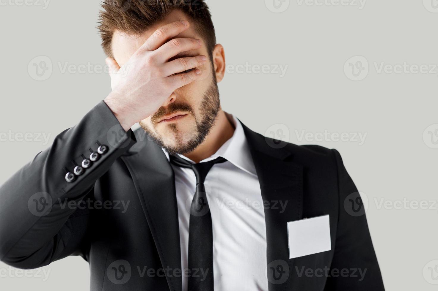 Failed management. Frustrated young man in formalwear covering face with hand while standing against grey background photo