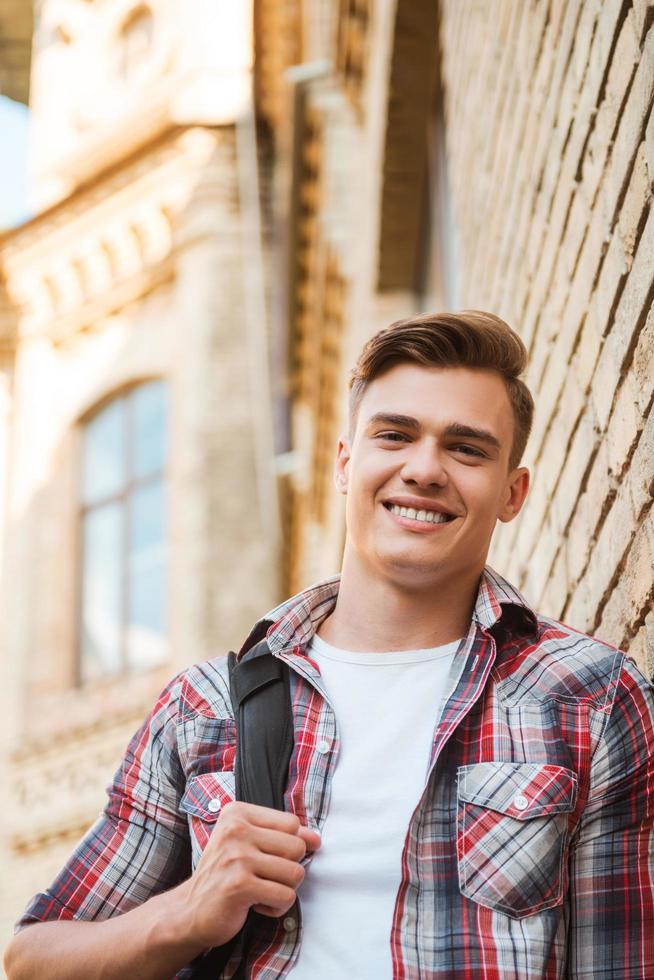 estudiante feliz vista de ángulo bajo de un joven apuesto que lleva una mochila en un hombro y sonríe mientras se apoya en la pared de ladrillo foto