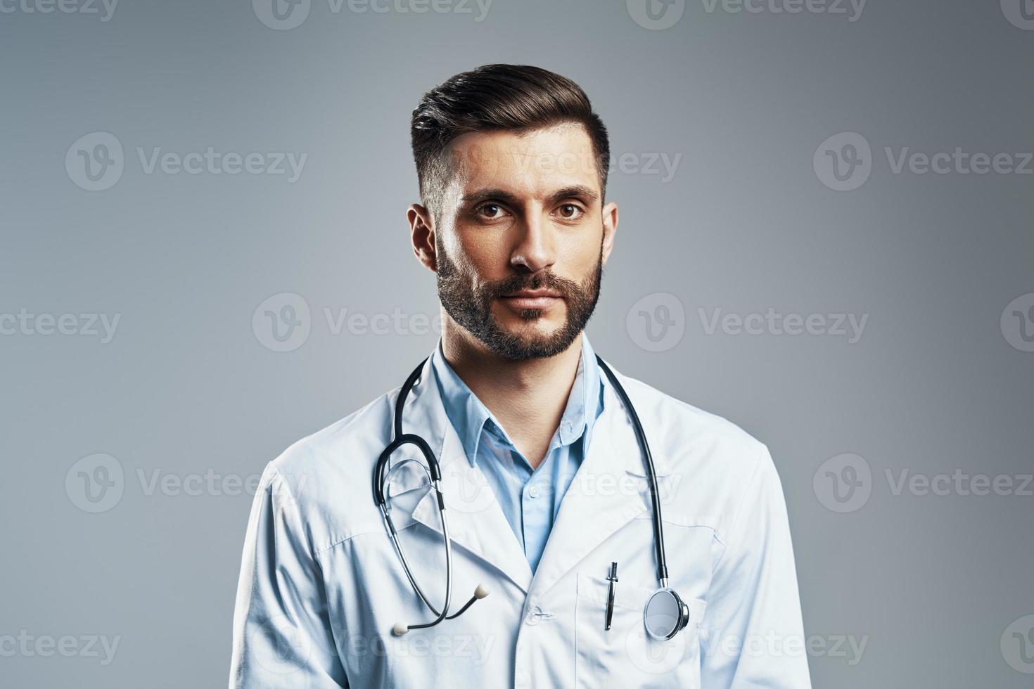 Handsome young man in white lab coat looking at camera while standing against grey background photo