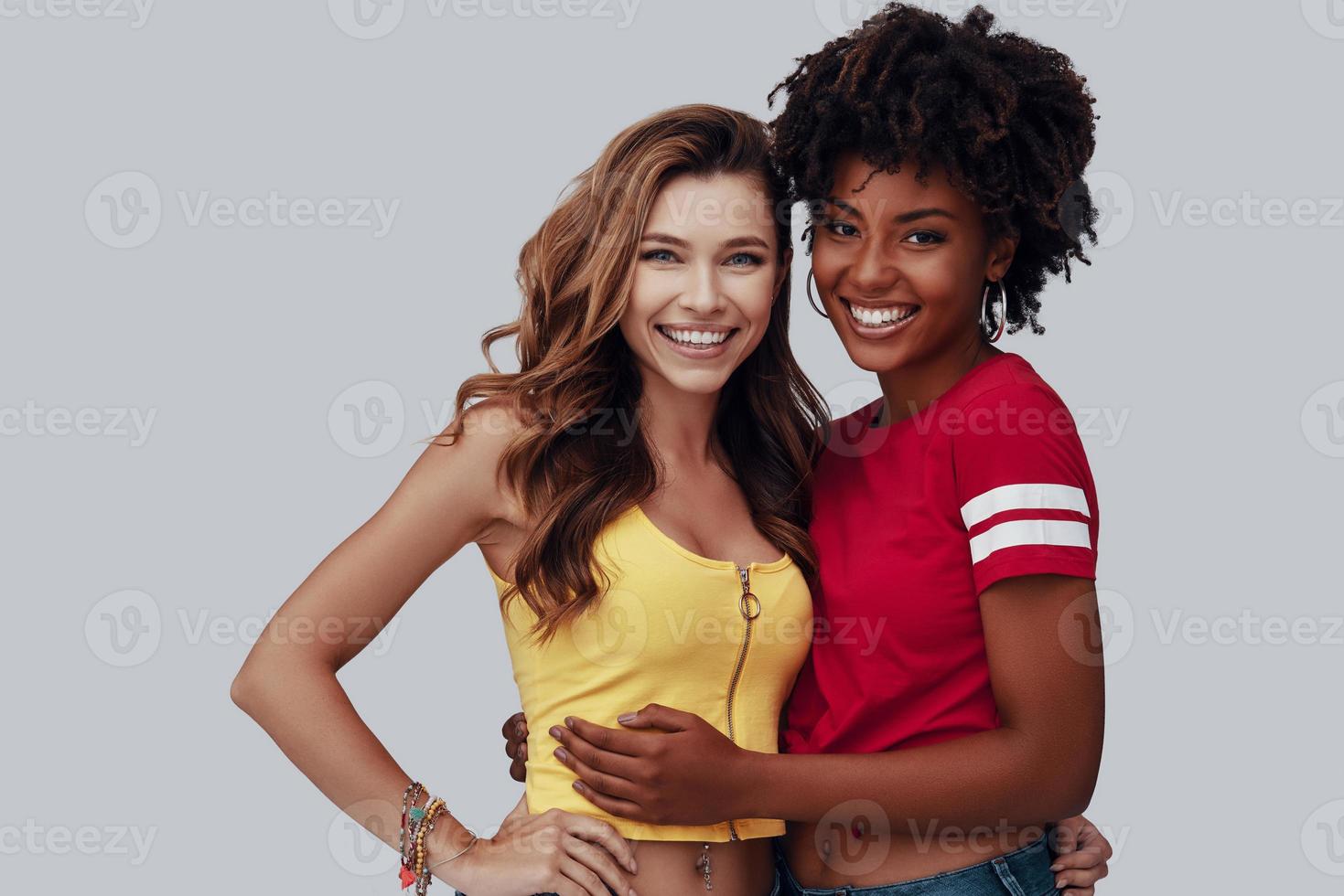 Two attractive young women looking at camera and smiling while standing against grey background photo