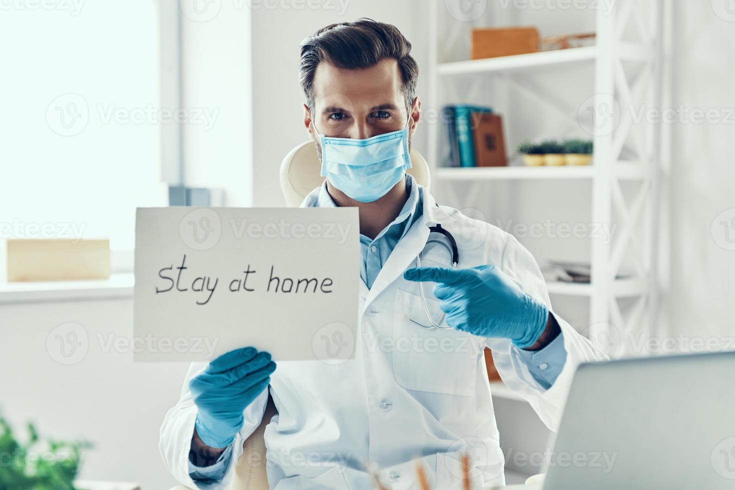 Young man in white lab coat and protective mask showing stay at home saying written on the paper while sitting indoors photo