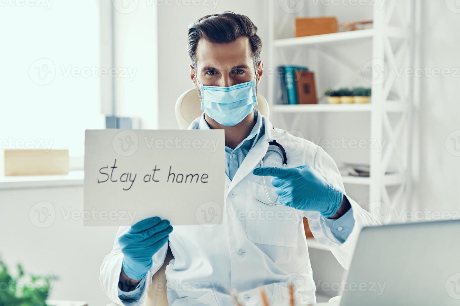 joven con bata blanca de laboratorio y máscara protectora que muestra quedarse en casa diciendo escrito en el papel mientras está sentado en el interior foto