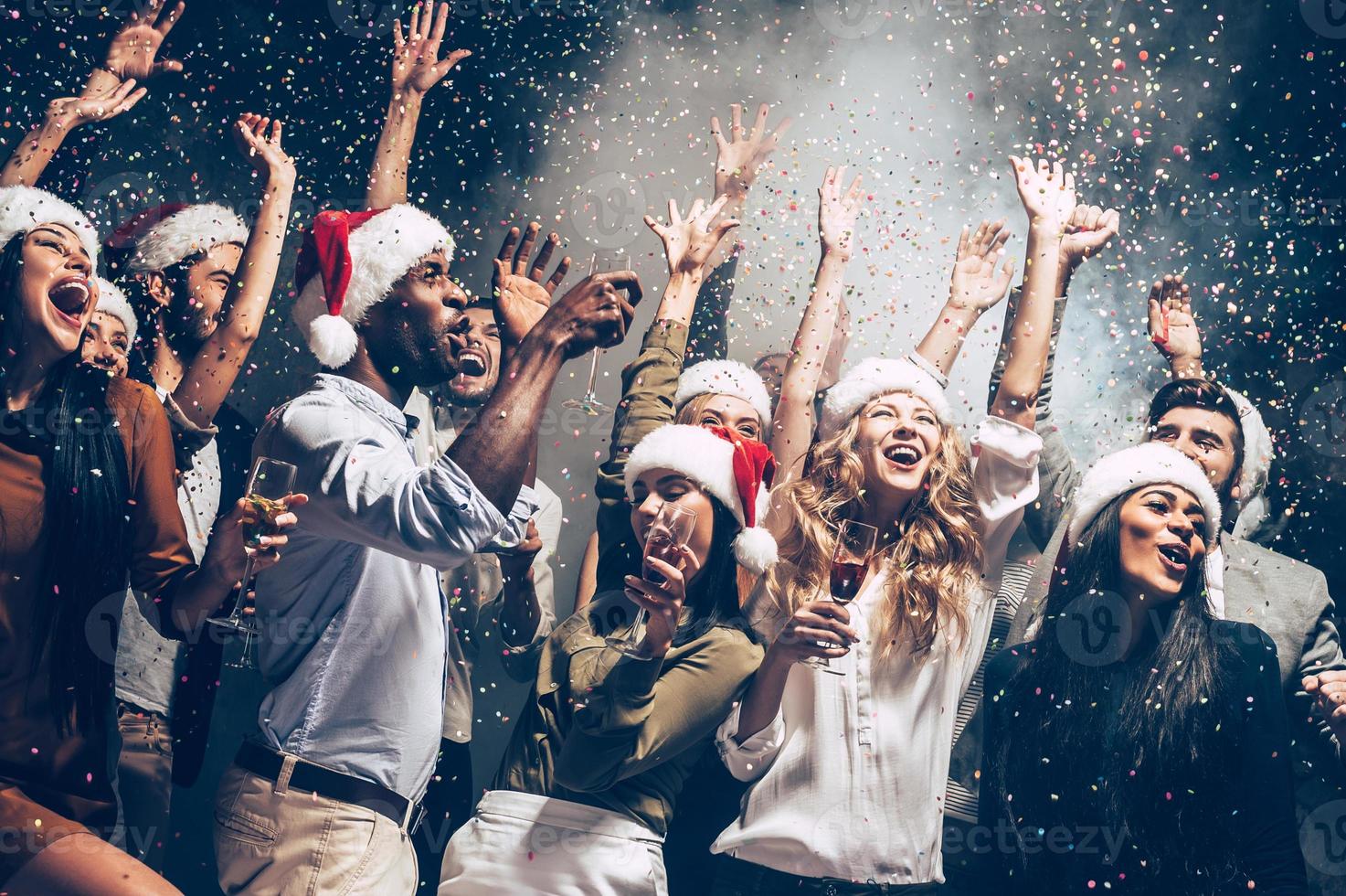 They love Christmas. Group of beautiful young people in Santa hats throwing colorful confetti and looking happy photo