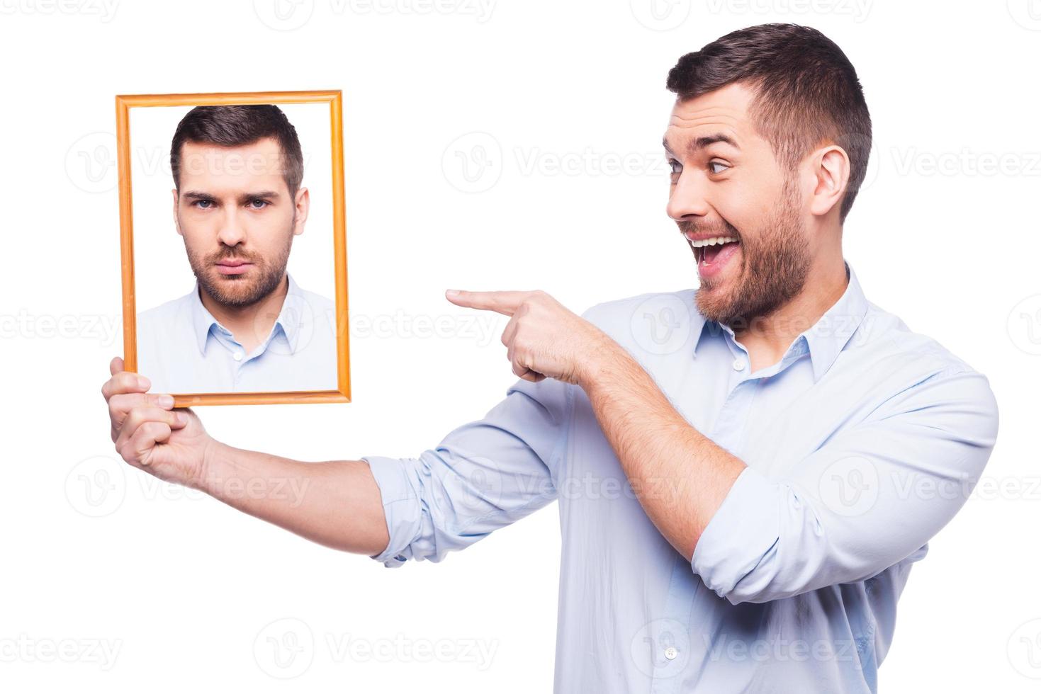 Give me a smile Handsome young man in shirt holding a picture of himself with upset face and pointing it with smile while standing against white background photo