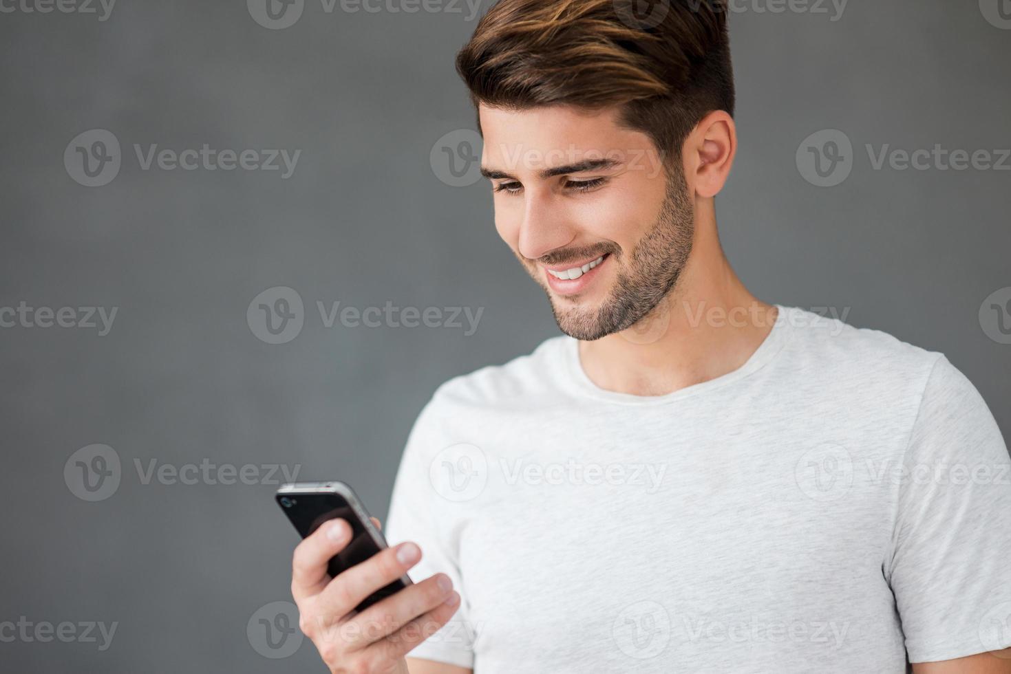 Texting to friend. Happy young man holding mobile phone and smiling while standing against grey background photo