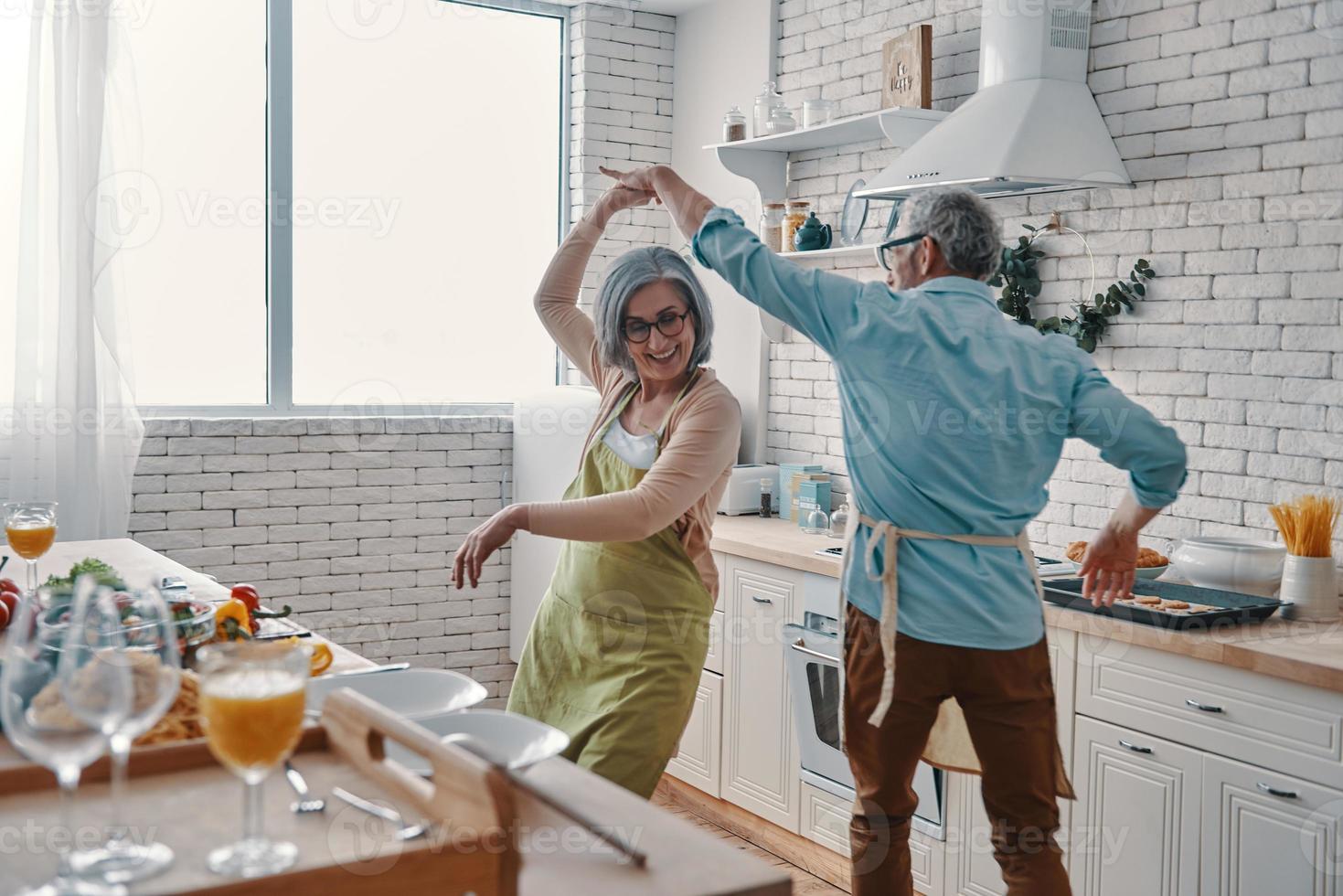 Beautiful playful senior couple in aprons dancing and smiling while preparing healthy dinner at home photo