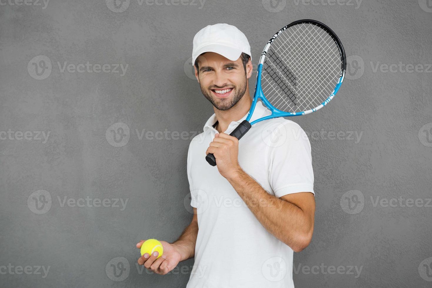 This game is more than hobby Happy young man in sports clothes carrying tennis racket on his shoulder and smiling while standing against grey background photo