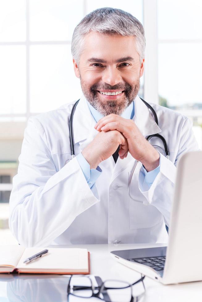 siempre dispuesto a ayudar. un médico de pelo gris maduro y confiado que se inclina la cara en las manos y sonríe mientras se sienta en su lugar de trabajo foto