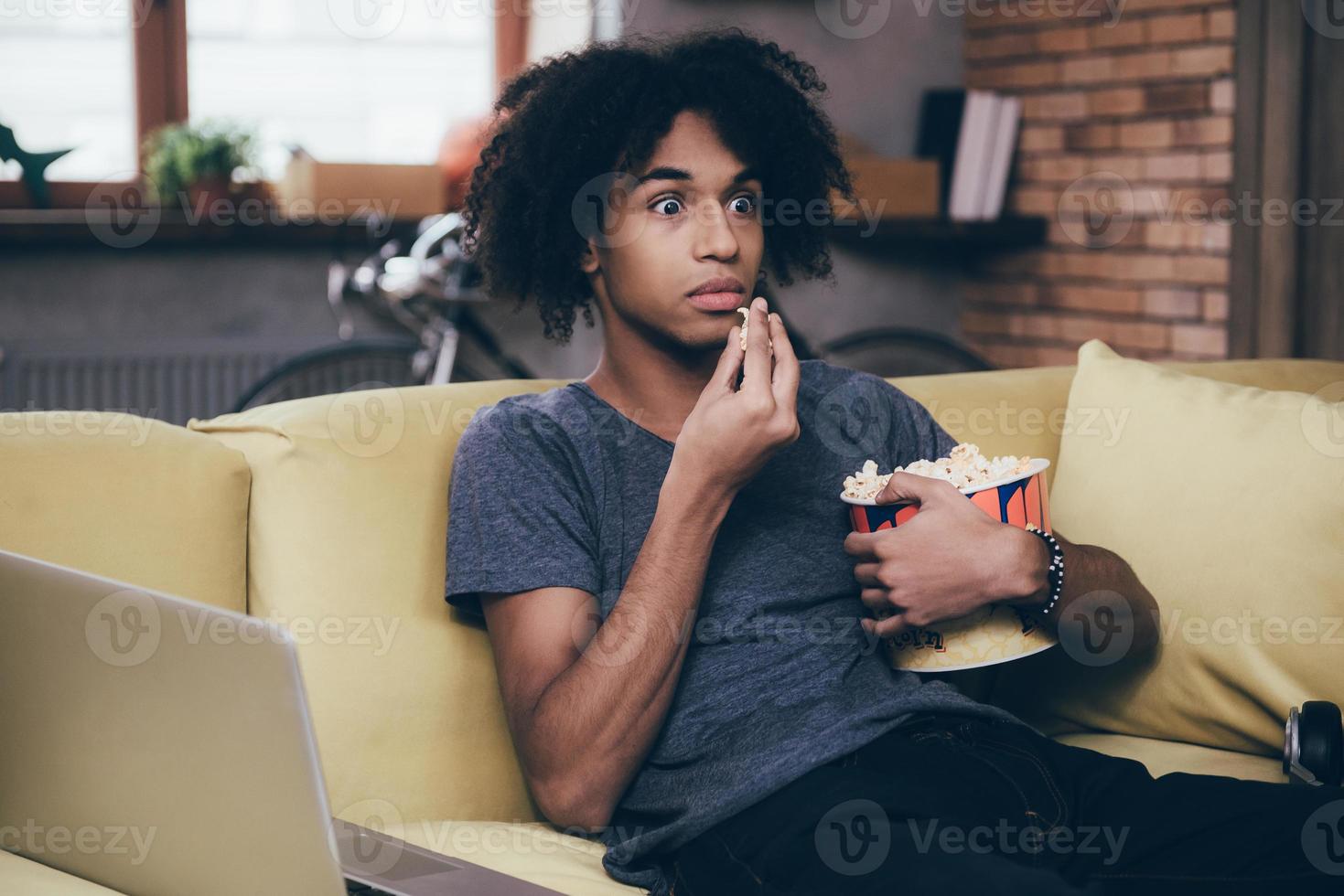 Horror movie. Young African man watching TV and looking scared while eating popcorn sitting on the couch at home photo