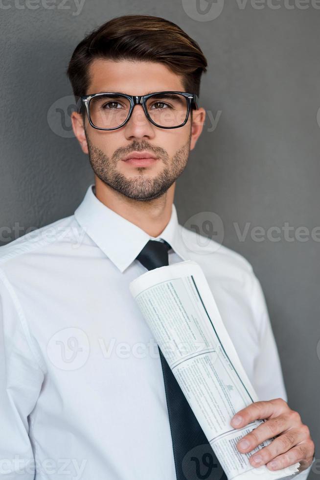 Thinking about new solutions. Confident young man in shirt and tie holding newspaper and looking away while standing against grey background photo