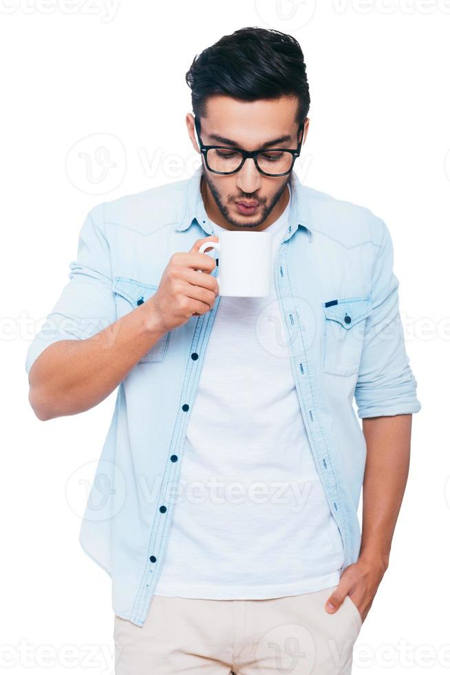 Too hot coffee. Handsome young Indian man holding cup with coffee and blowing on it while standing against white background photo