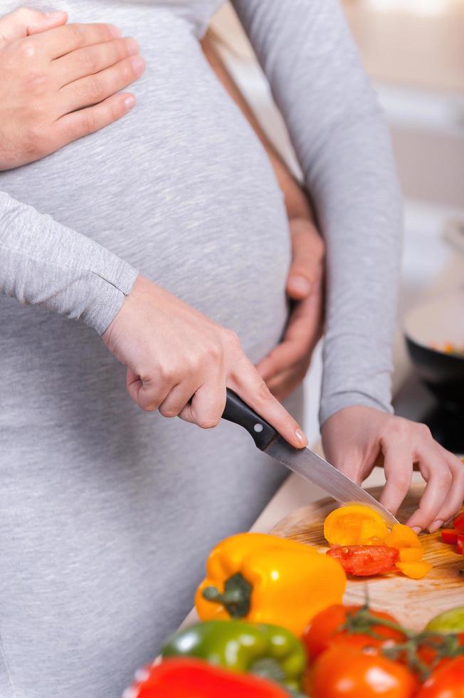 Only healthy food. Happy young couple cooking together in the kitchen while man touching abdomen of his pregnant wife photo