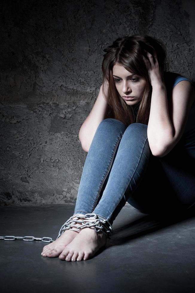 Feeling hopeless. Young woman trapped in chains holding head in hands while sitting on the floor in a dark room photo