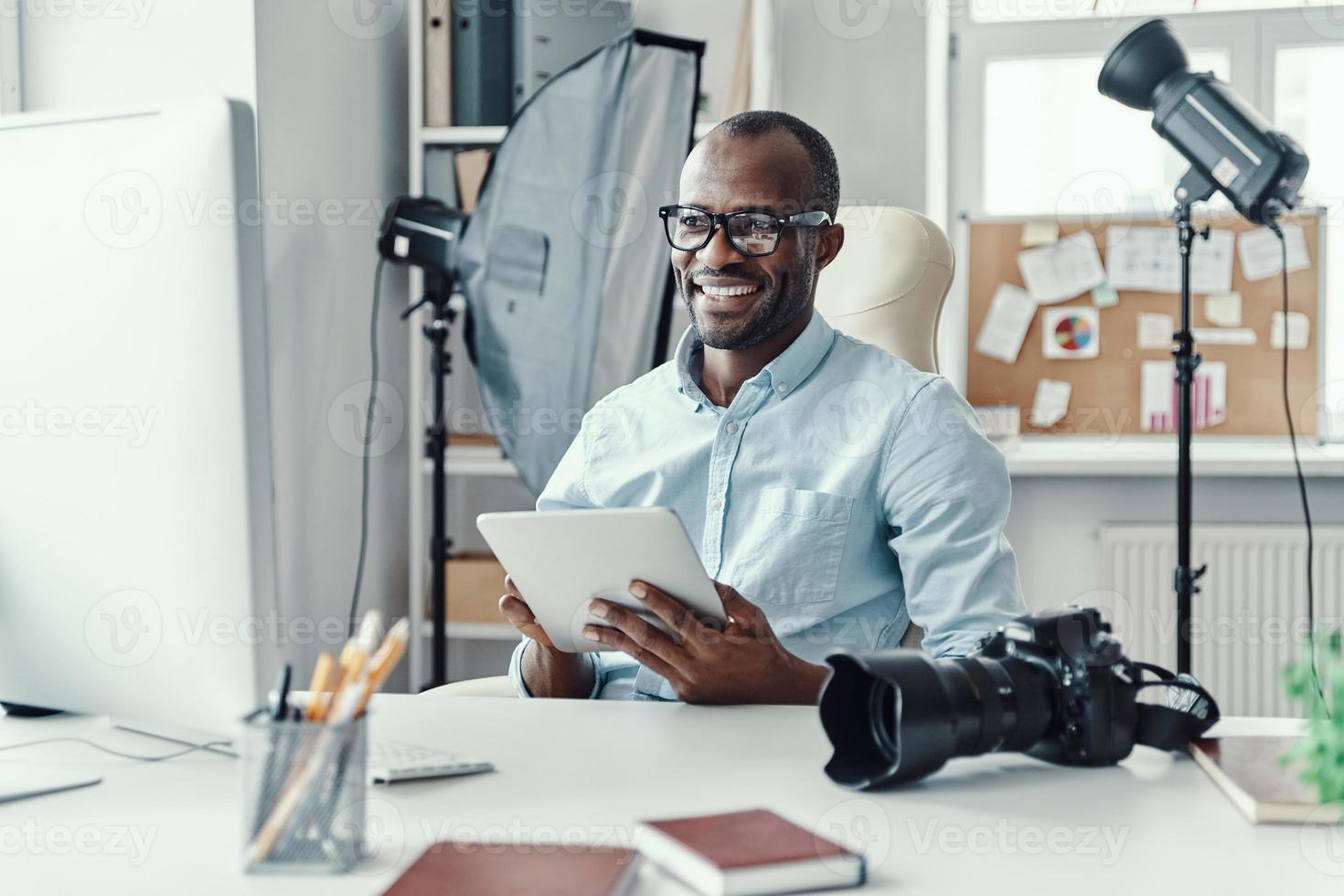 Happy young African man using digital tablet and smiling while working in the modern office photo