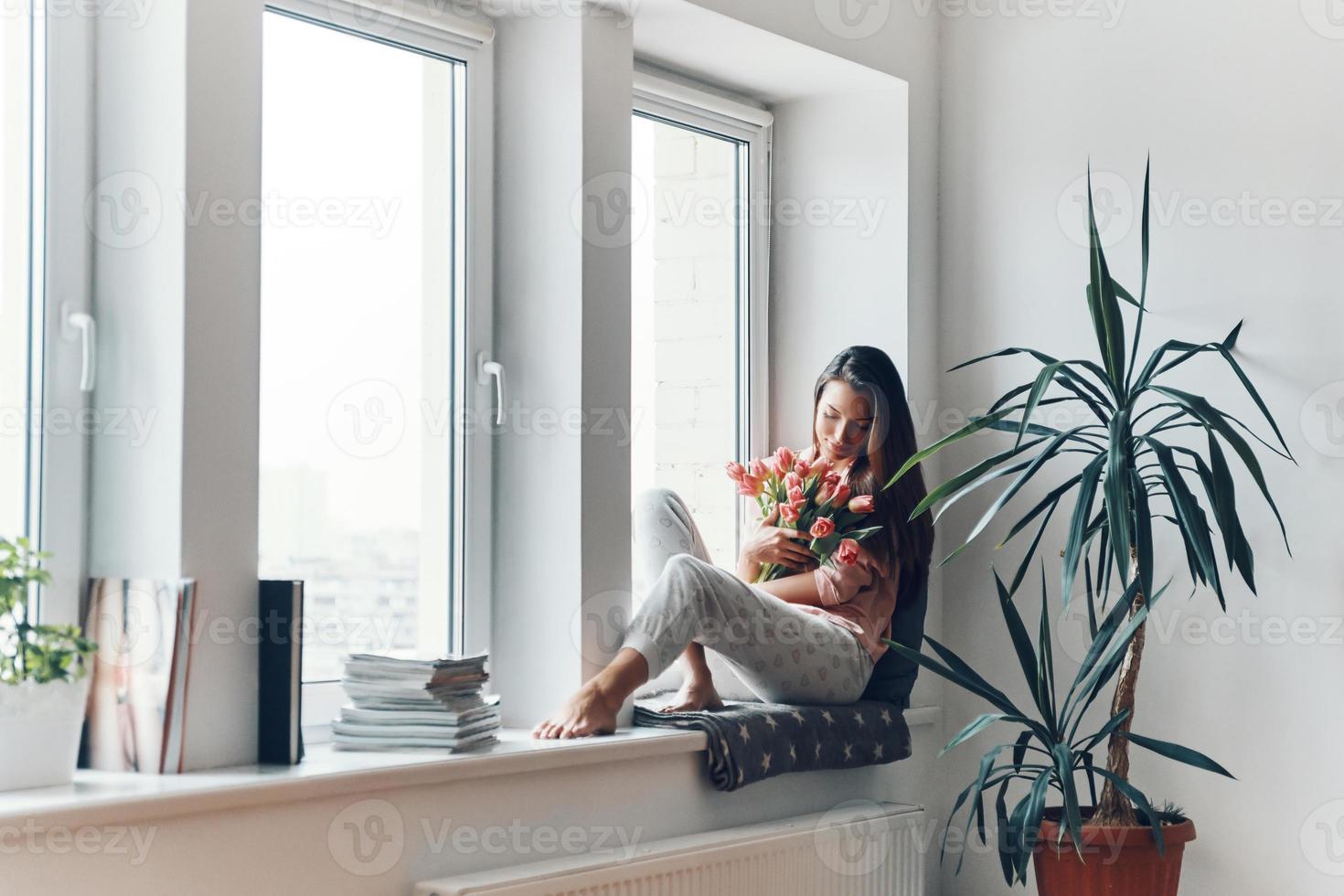 Beautiful young woman in cozy pajamas enjoying her bouquet of tulips while resting on the window sill at home photo
