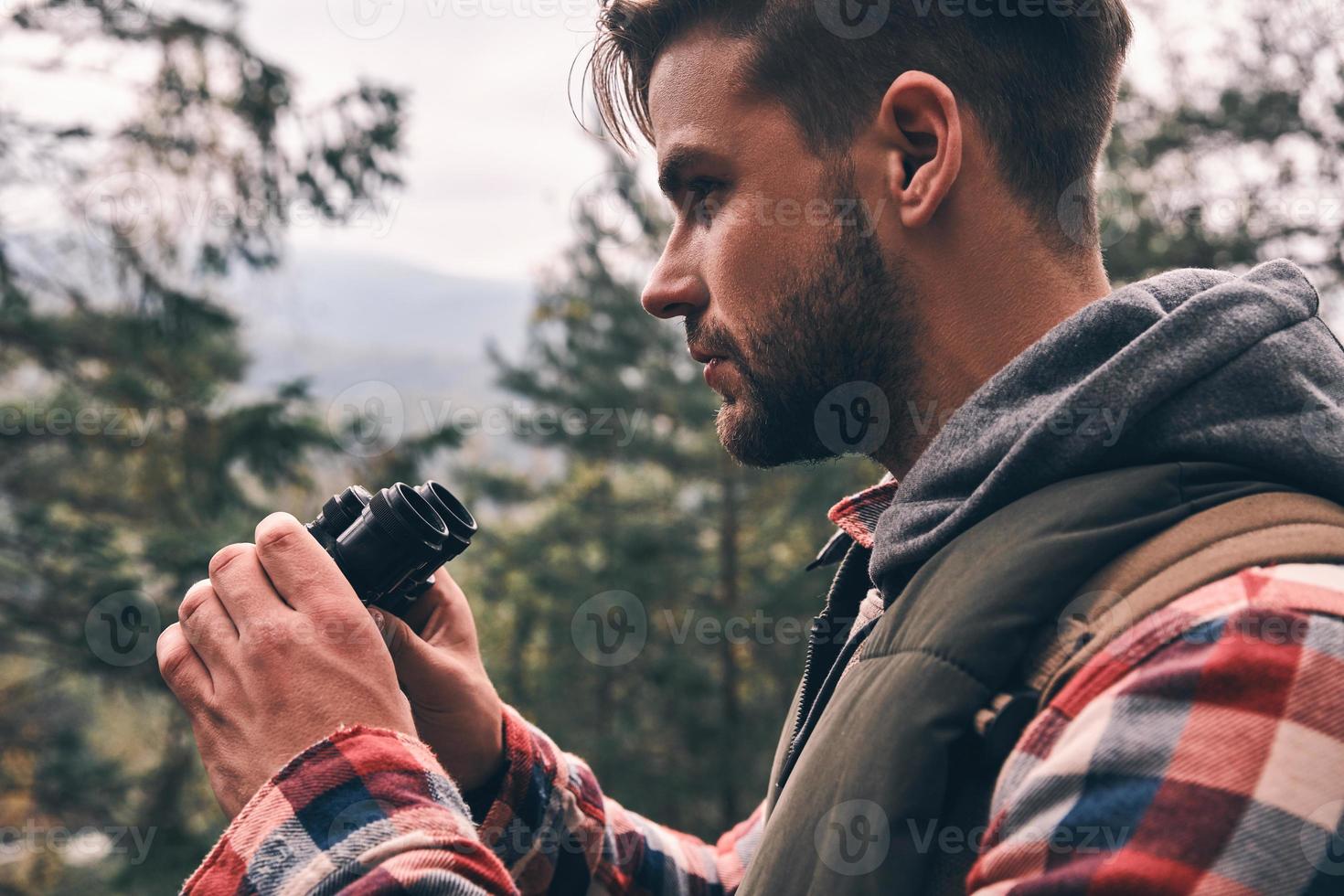 Handsome hiker. Modern young man with backpack looking away through binoculars while standing in the woods photo