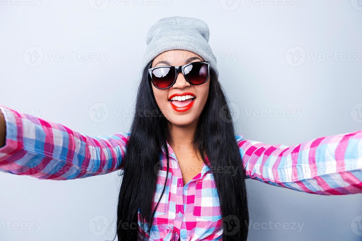Capturing fun. Happy young African woman in funky clothes making selfie while standing against grey background photo