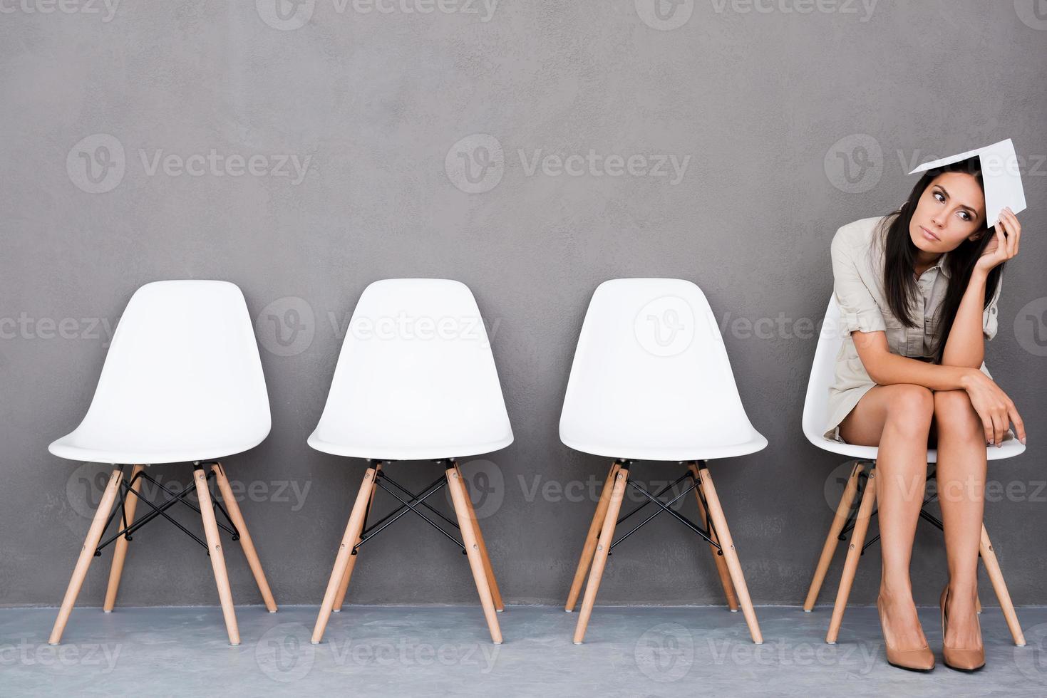 Feeling bored of waiting. Bored young businesswoman holding paper on her head and looking away while sitting on chair against grey background photo