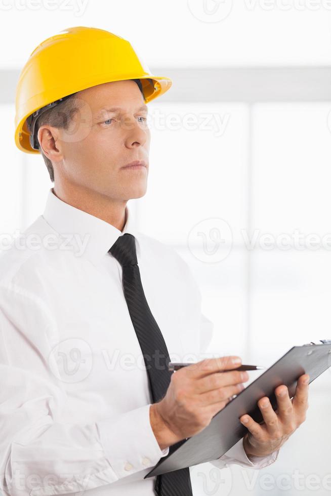 Construction foreman. Confident mature man in formalwear and hardhat writing something in his note pad and looking away photo