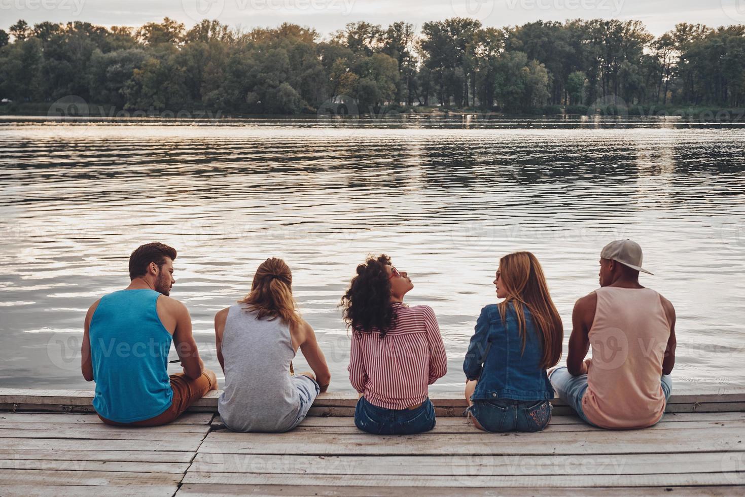 tiempo sin preocupaciones con amigos. vista trasera de jóvenes con ropa informal hablando mientras están sentados en el muelle foto