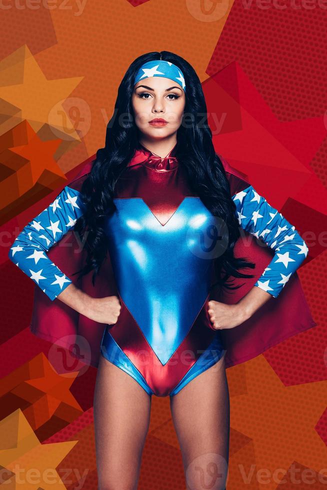 Evil can not handle with her. Beautiful young woman in superhero costume looking at camera and keeping hands on hips while standing against colorful background photo