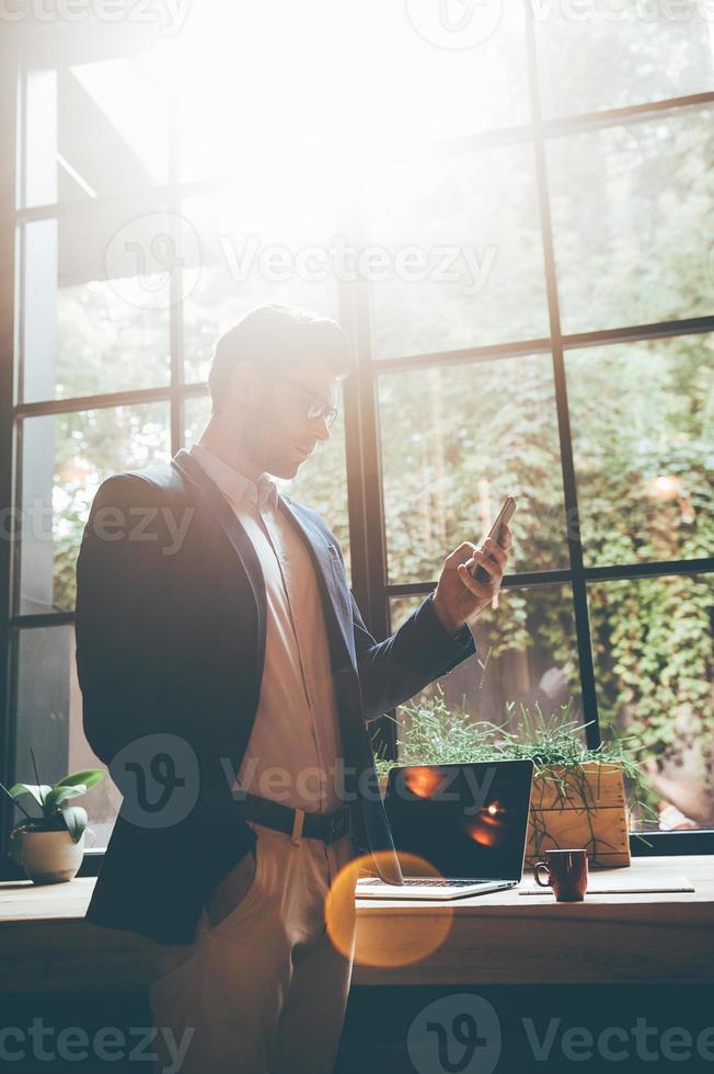 Typing quick business message. Low angle view of confident young man holding smart phone and looking at it while standing in front of the big window in office or cafe photo