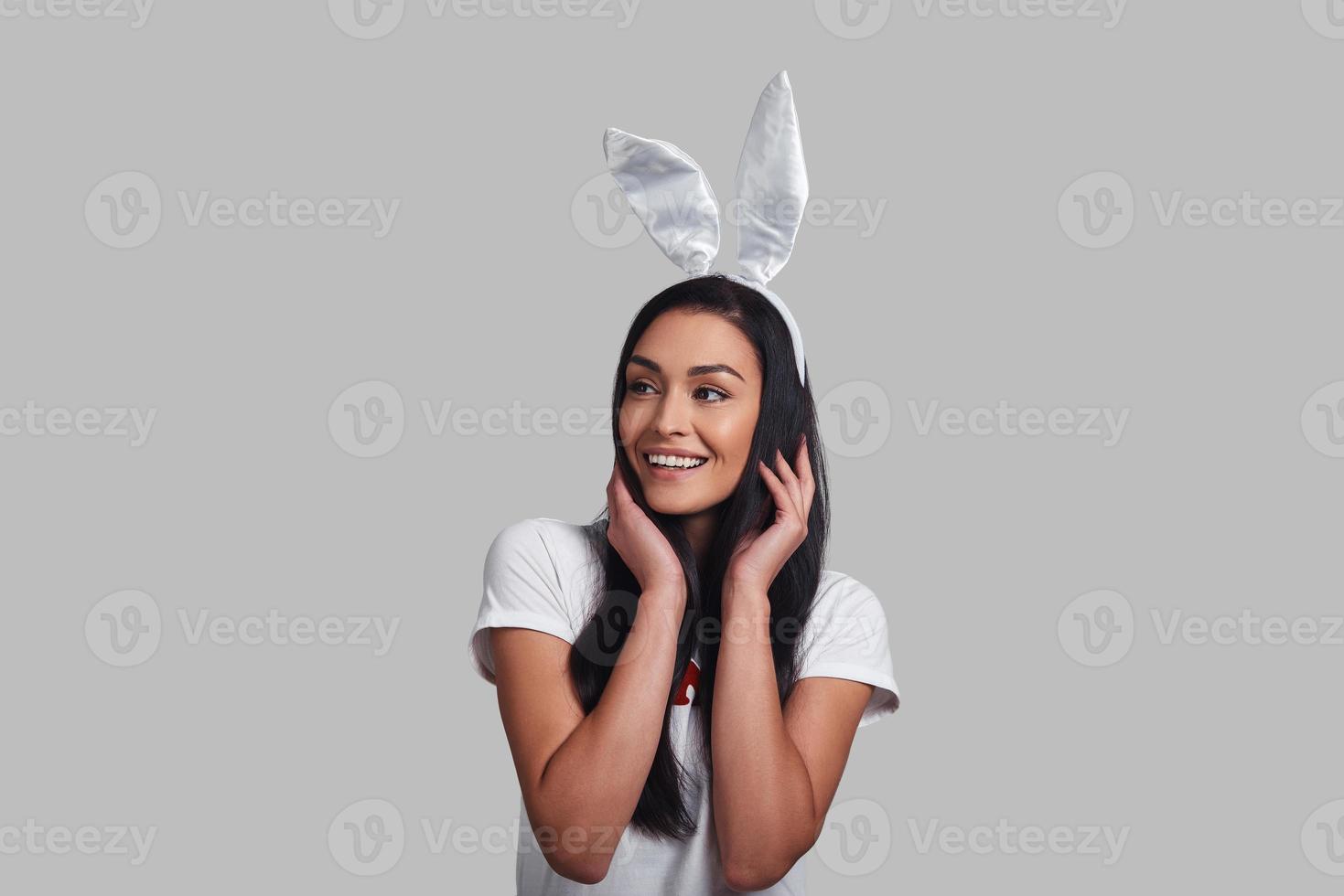 Cuter than any bunny. Attractive young woman in bunny ears looking away and smiling while standing against grey background photo