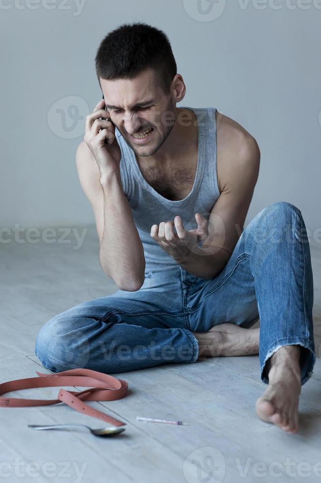 I need more Furious young man sitting on the floor and talking on the mobile phone while with syringe and spoon laying near him photo